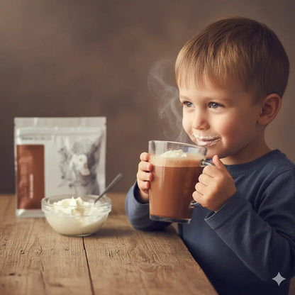 Egy szőke kisfiú tejcsokis forrócsokit iszik, tejszínhabbal egy asztalon. A szája tejszínhabos. 